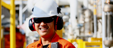 Man in orange work clothes, ear muffs and helmet stands in front of energy facility