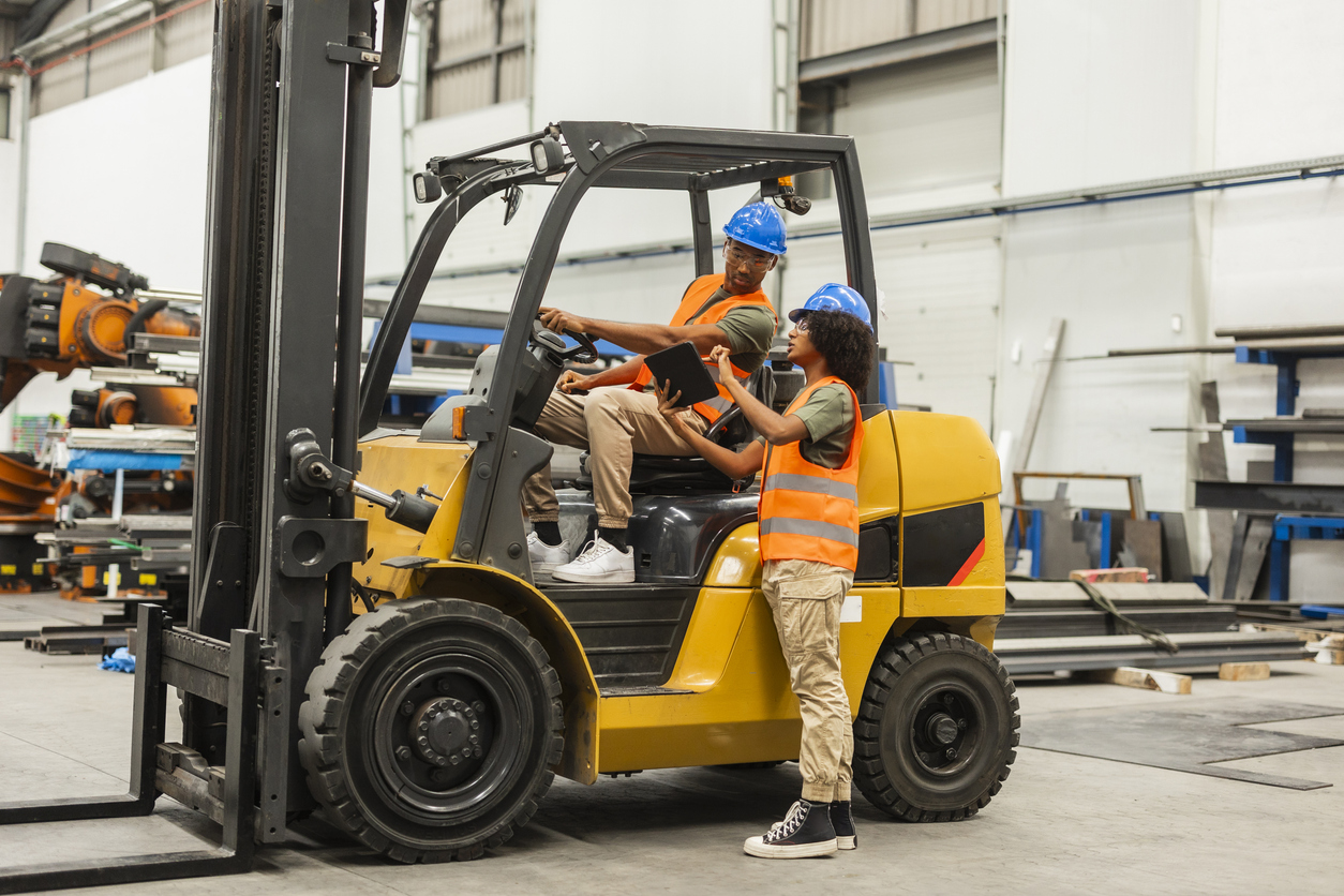 Two warehouse workers look at a tablet while one drives a forklift