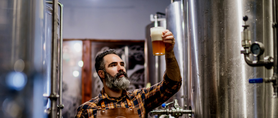 Brewer samples beer next to production equipment | Small Business