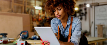 Lady leans over work desk using a tablet with tools on table behind her