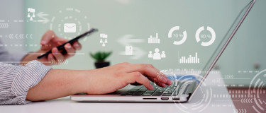 Hand types on laptop while holding a mobile phone in other hand with analytics graphics coming off the screen