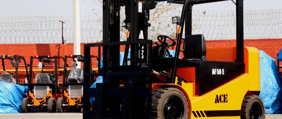 Yellow and black electric forklift parked on a concrete yard, with a row of further lift trucks and tarp-covered machines lined up behind it