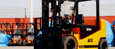 Yellow and black electric forklift parked on a concrete yard, with a row of further lift trucks and tarp-covered machines lined up behind it
