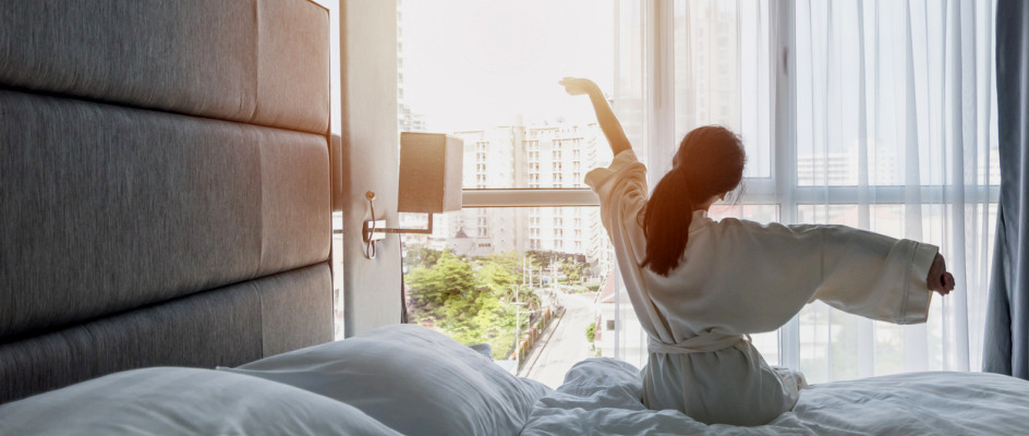 Hotel guest sits on bed with white linen looking out window stretching arms