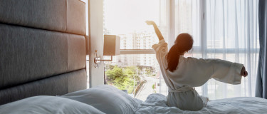 Hotel guest sits on bed with white linen looking out window stretching arms