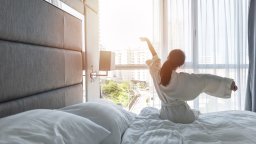 Hotel guest sits on bed with white linen looking out window stretching arms