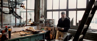 Man in a dark hooded jacket stands at a green workbench in a sunlit workshop, reading a tablet, surrounded by hand tools, small parts drawers and metal frames
