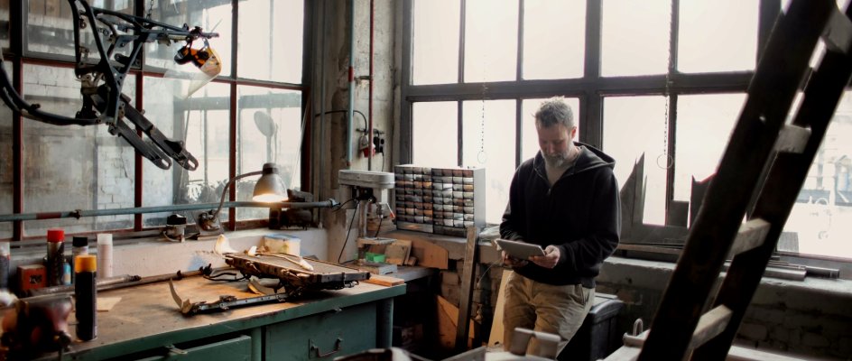 Man in a dark hooded jacket stands at a green workbench in a sunlit workshop, reading a tablet, surrounded by hand tools, small parts drawers and metal frames