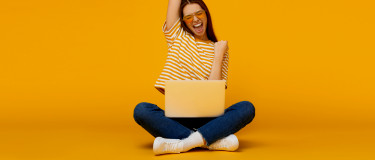 Woman sitting with laptop on legs celebrating success with yellow background