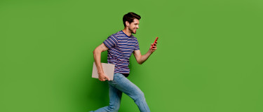 man with a laptop under the arm and a mobile phone in the hand jumps in the air with green background