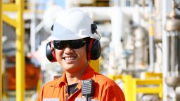 Man in orange work clothes, ear muffs and helmet stands in front of energy facility
